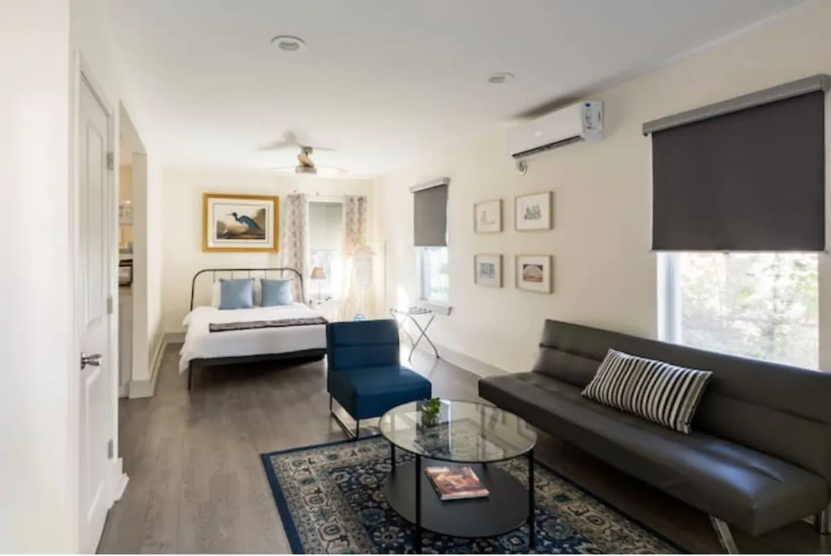 A serene bedroom and sitting area with a bed adorned with blue pillows, a modern blue chair, a glass coffee table with a book, and wall art. Natural light fills the space, creating an inviting atmosphere ideal for unwinding.