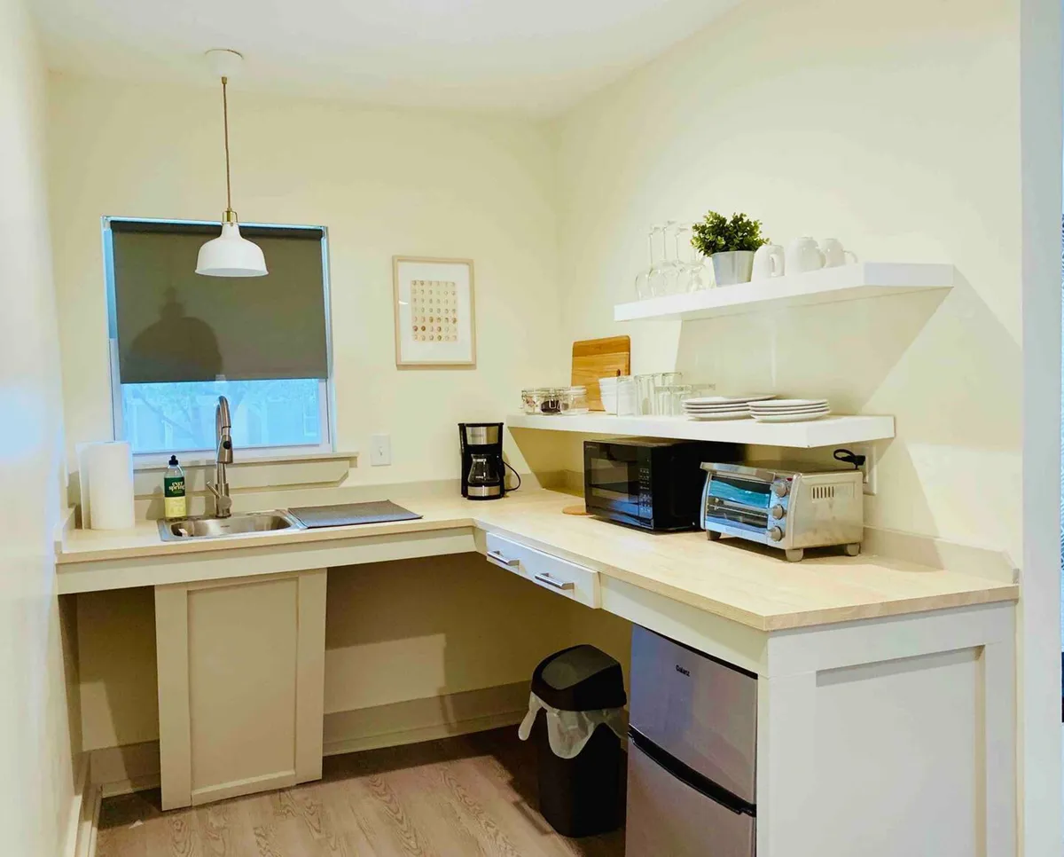 A cozy kitchenette with a sink, coffee machine, toaster oven, and open shelves neatly stocked with dishes. The natural light by the window enhances the inviting atmosphere, and the setup is ideal for preparing simple meals or enjoying a quiet coffee moment.