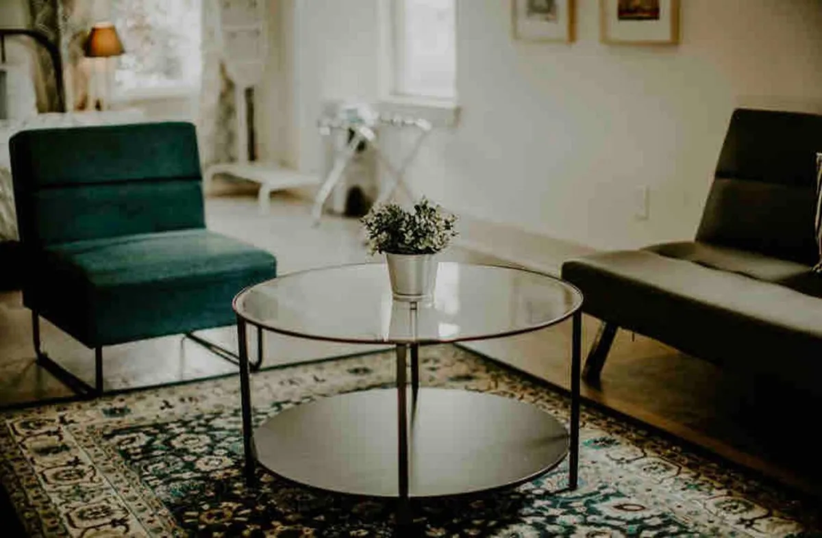 A tastefully designed living area featuring a green armchair, a plush gray sofa, a round glass coffee table with a small potted plant, and a decorative rug. The arrangement is inviting, suggesting a welcoming space for relaxation or socializing.