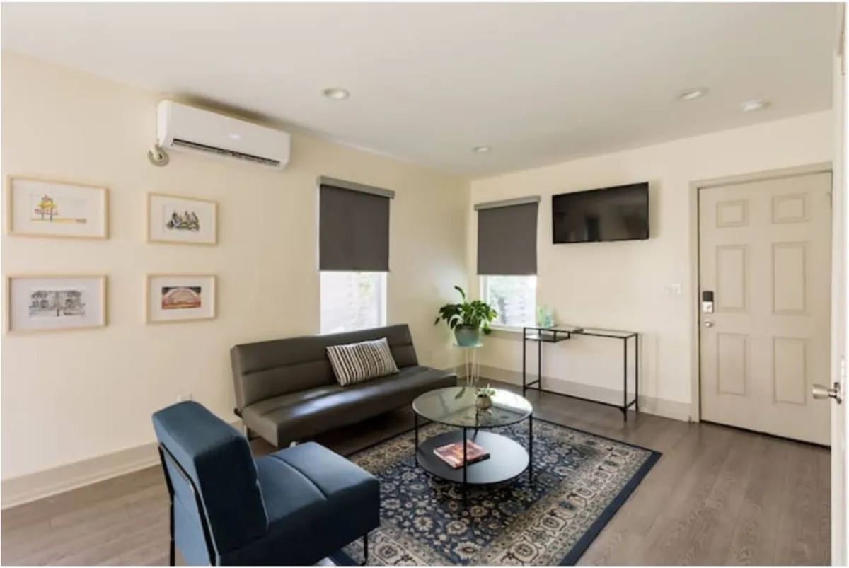 A bright, comfortable living room featuring a modern sofa, sleek coffee table, inviting houseplant, large windows letting in natural light, and stylish artwork on the wall. The setup hints at a space designed for relaxation, socializing, or enjoying entertainment, with a direct view of the living area amenities.