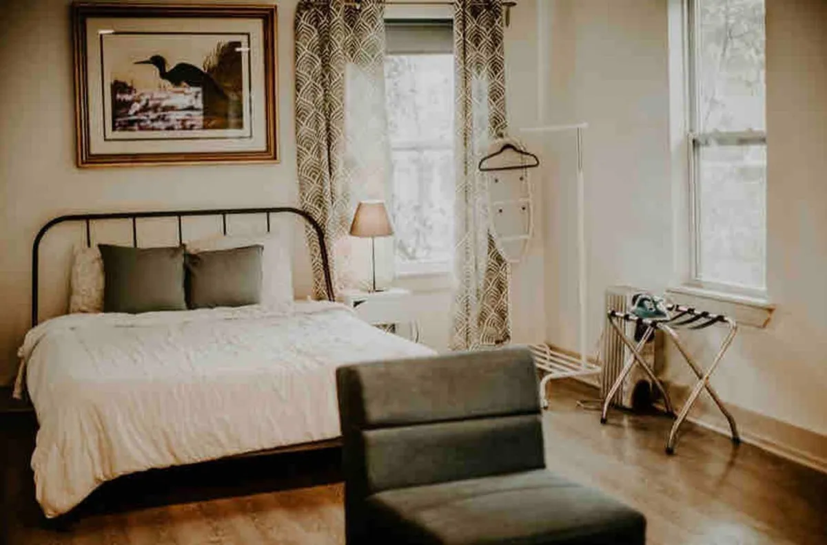 The image showcases a cozy, sunlit bedroom featuring a plush bed, inviting soft linens, artfully arranged framed artwork above the headboard, a stylish lamp casting warm light, and a window that bathes the space in natural morning glow. The ambiance is peaceful and thoughtfully curated, ideal for relaxation or quiet moments with a book or coffee.