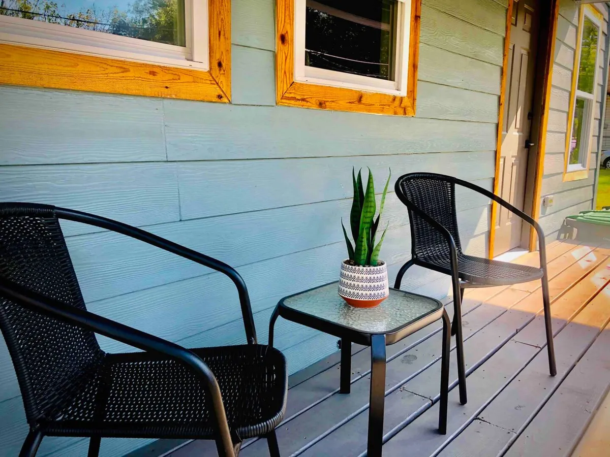 A shaded backyard seating area featuring two black chairs, a small glass table, and a potted plant. The space is tranquil and designed for relaxation, perfect for enjoying morning coffee or unwinding at sunset.
