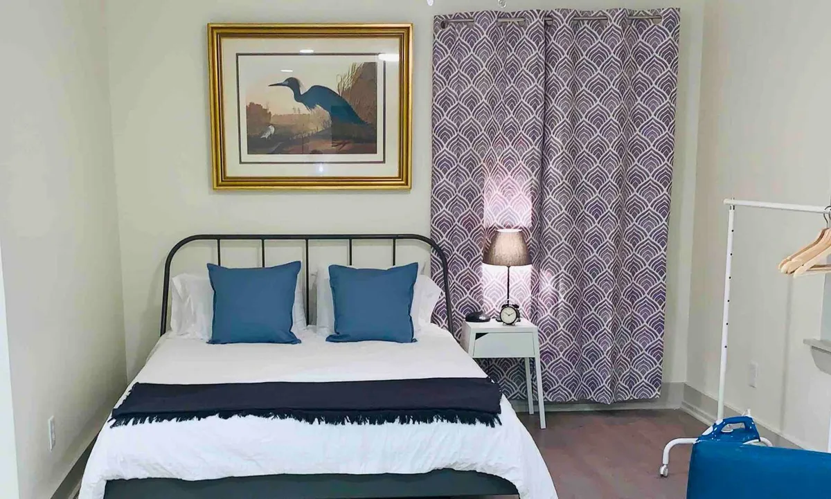 A cozy bedroom scene featuring a plush bed with quality linens, framed artwork above the headboard, soft curtains filtering natural light, a lamp on the nightstand, and a warm, tranquil atmosphere ideal for relaxation and restful sleep.