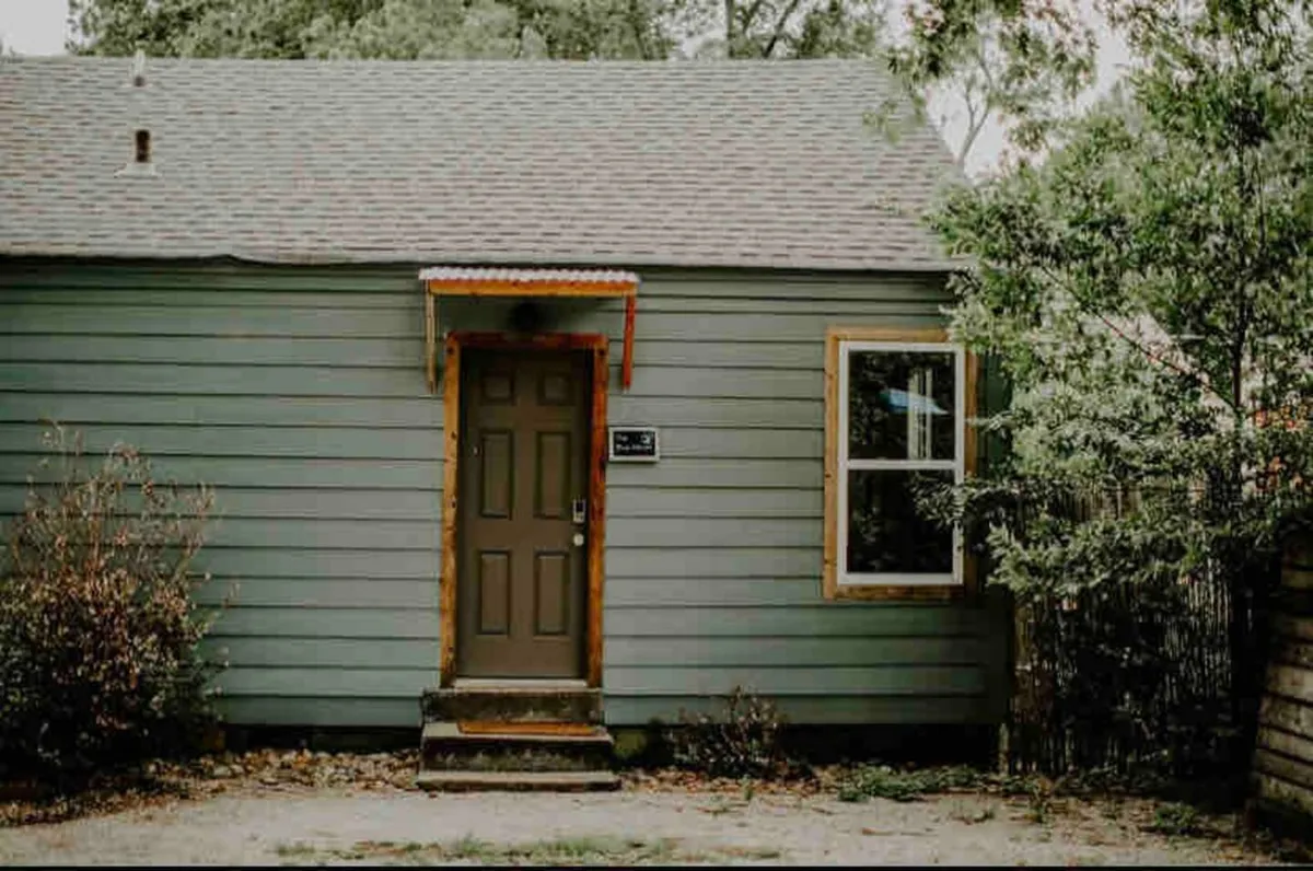 The image highlights the home's front door and window, bordered by a landscaped area and a porch. The greenery and meticulous upkeep signal a tranquil and private setting, ideal for solo travelers seeking both comfort and security.